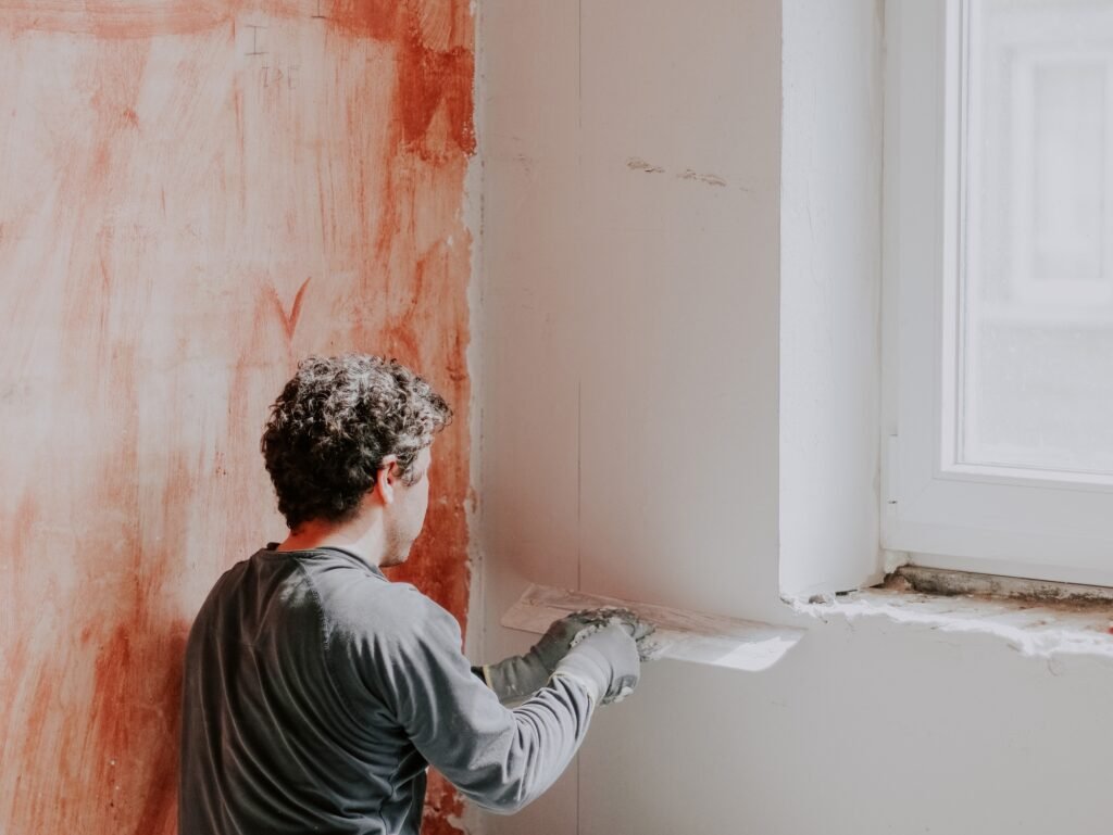 A young caucasian plasterer holds a large trowel and levels a freshly plastered wall